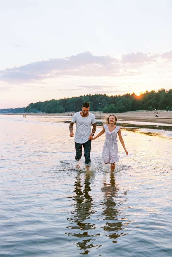 A joyful couple holding hands and walking through the shallow waters at sunset, reflecting happiness and romance.